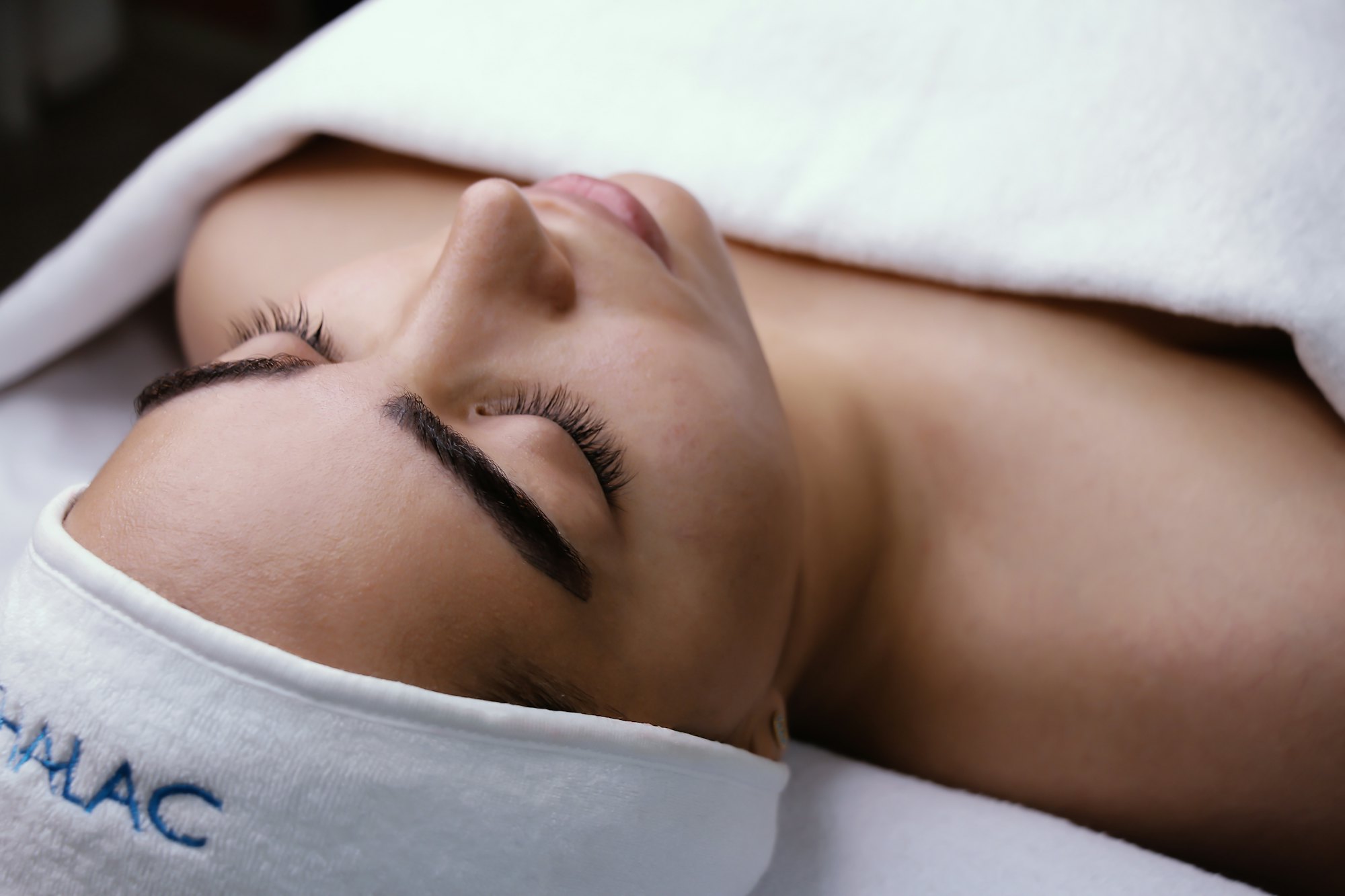 Client resting with eyes closed during a facial, hair wrapped in a white towel